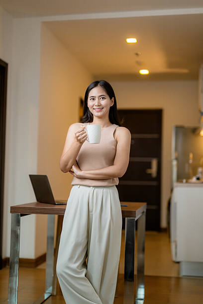 Portrait of a smiling young Asian businesswoman drinking coffee while working from home, standing next to her desk with a laptop on it