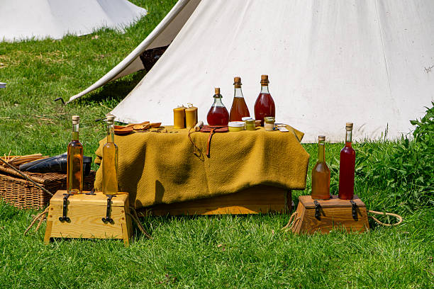 Historical reenactment from the Middle Ages or the Renaissance. A camp table covered with a yellow tablecloth, on which bottles of homemade liqueurs or wine, clay cups and kitchen accessories are placed. In the background, a white tent and green grass are visible, suggesting an outdoor festival or historical reenactment. The perfect photo to illustrate a historical, ethnographic or culinary theme.