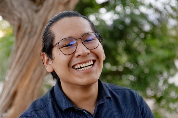 istock Cheerful young man with glasses laughing, blurred green background, tree trunk partially visible in natural setting 2213386320