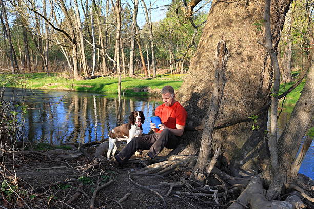 Middle-aged man rests against a tree by the stream to read a book with his dog