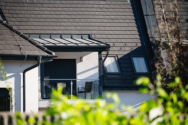 A sleek modern balcony with glass railing and outdoor furniture is part of a white house with a sharply angled black tiled roof. The foreground greenery and sunlight create a serene suburban ambiance.