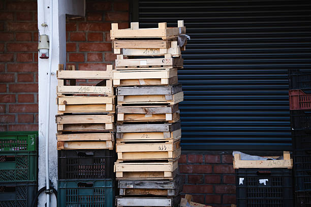 A tall stack of weathered wooden crates stands beside plastic bins against a brick wall and closed metal shutter, likely at a market or store. The rough textures and utilitarian setting evoke a sense of everyday commerce and manual labor.