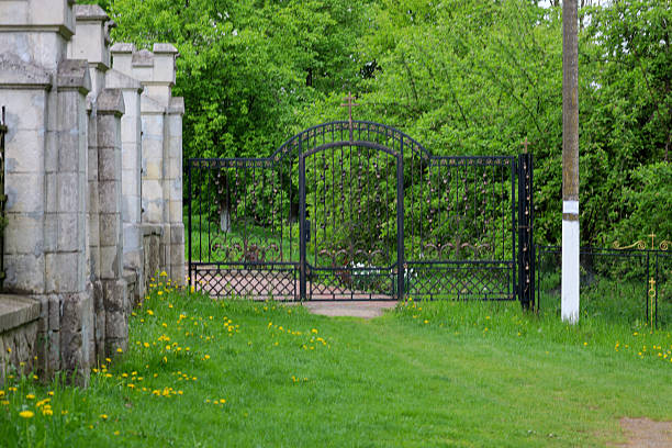 istock A black wrought iron gate with decorative elements is closed, surrounded by a green lawn with yellow dandelions and a stone fence against a background of green trees. 2213434797