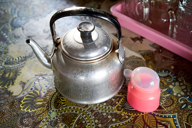 A classic aluminum teapot, a staple in many households for brewing tea or coffee, sits on a vibrant, ethnically patterned tablecloth alongside a simple toothpick holder. This scene evokes a sense of tradition, daily rituals of beverage preparation, or a cozy, homey atmosphere often found in Southeast Asian settings.