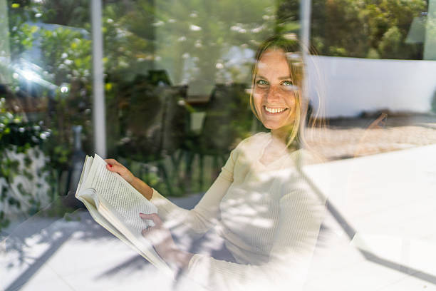 istock Young woman smiling by the window reading a book 2213452831