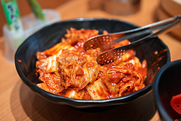 A close-up of spicy Korean kimchi served in a black bowl with metal tongs, placed on a wooden dining table.