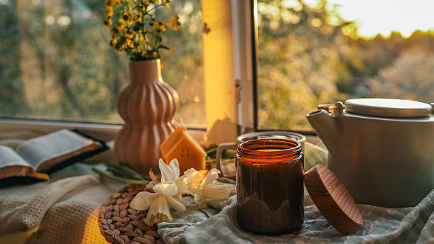 Peaceful spring still life with a teapot, candle jar, flowers, and open book on a windowsill. Golden hour light creates a warm and cozy ambiance with soft natural textures