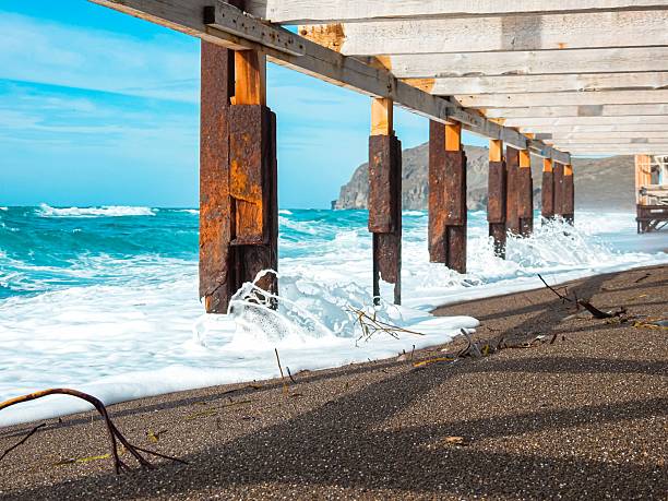 Waves crashing against a wooden pier on a sandy beach with clear blue skies and distant mountains.