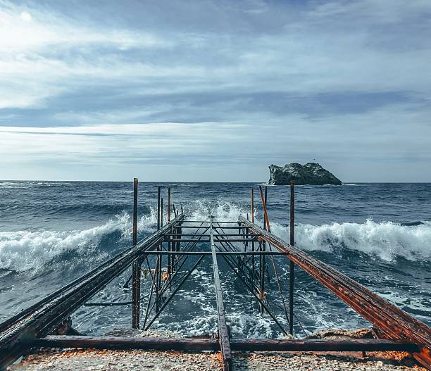 Rusty pier extending into the ocean with waves crashing against it, under a cloudy sky, and a distant island visible on the horizon.