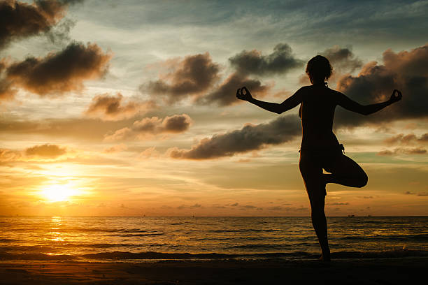 istock A woman practicing yoga while standing on a tropical beach during a stunning sunset, surrounded by serene beauty and warm light. 2213480437