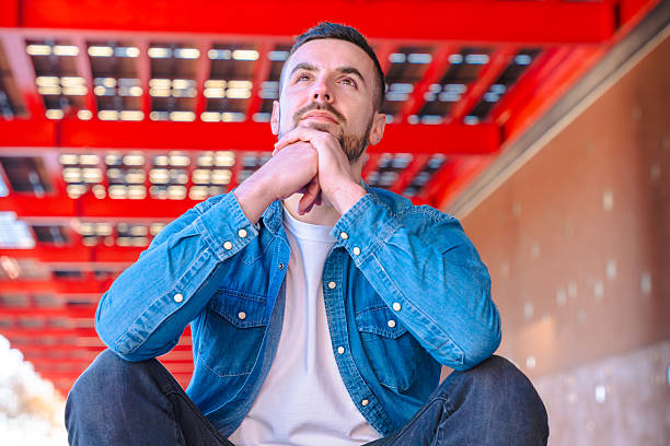 Thoughtful man in denim shirt sitting under a striking red urban structure, conveying reflection, lifestyle, and personal contemplation in a modern city environment with strong geometric contrast