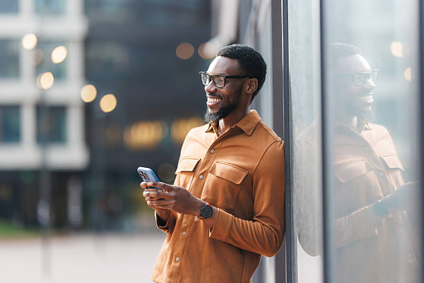 Man leaning against reflective glass wall, smiling and using smart phone