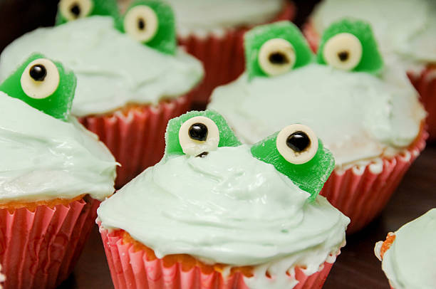 Close-up of charming frog-themed cupcakes perfect for a children's birthday party or spring celebration. Each cupcake features white frosting with decorative green candy frog faces peeking out, complete with expressive round candy eyes with black pupils. The cupcakes are served in bright pink paper liners, creating a playful contrast with the white frosting and green frog elements. These whimsical treats would make an eye-catching centerpiece for any kid-friendly celebration or themed event.