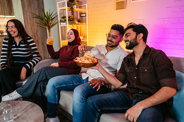 Group Of Indian And Muslim Friends Watching A Funny Movie Together On The Sofa While Eating Chips Group of Indian and Muslim friends watching a funny movie together on the sofa while eating chips. family eating potato chips stock pictures, royalty-free photos & images