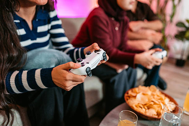 Young woman playing video games on a joystick with friends on the sofa while eating chips. Close-up.