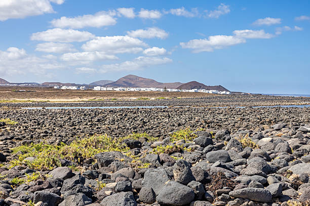 This image presents a striking view of La Santa, a coastal town on Lanzarote, seen across the island’s rugged volcanic and arid landscape. The foreground features dark, rocky terrain, shaped by past eruptions, with sparse vegetation adding texture to the barren environment. In the distance, the white buildings of La Santa contrast sharply against the brown hills and clear blue sky, emphasizing the town’s isolation within the dramatic scenery. The image highlights Lanzarote’s unique geology and the harmonious balance between human settlement and its stark natural surroundings.