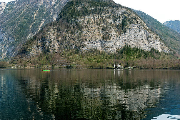 Tranquil alpine lake with crystal clear water reflecting majestic mountain. Small yellow boat adds vibrant accent to serene landscape. Rocky cliffs covered with pine trees