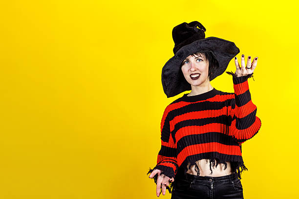 istock Halloween Theme. Young witch in striped sweater and black hat making magic or spell against yellow background. Free space for your text 2213551106