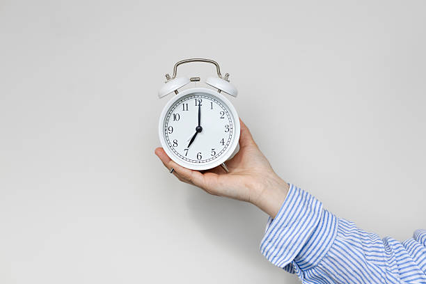 istock Woman hand holding white alarm clock on white background. Seven o'clock. 2213552244