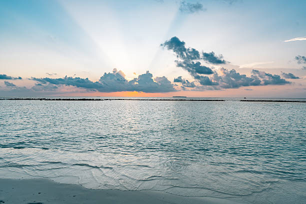 Scenic view of a Maldives beach at sunset with sunbeams shining through clouds and calm ocean water