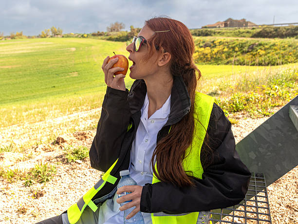 Female engineer in a safety vest taking a break outdoors, enjoying an apple and hydrating with water while sitting on a metal structure near an industrial site