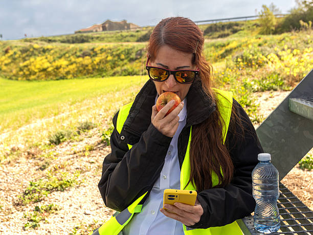 Female engineer enjoying a well-deserved break, savoring a nutritious apple while checking her smartphone. Balancing work and wellness in a serene outdoor setting