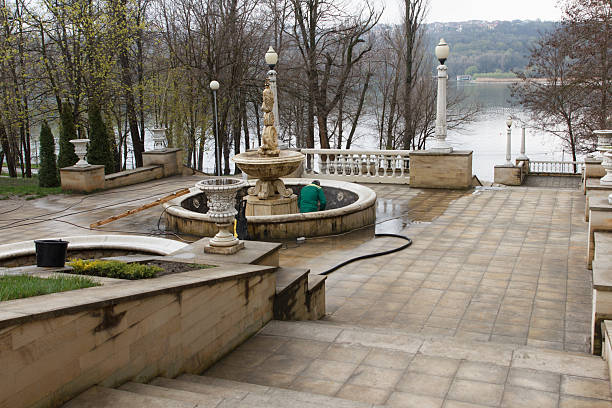 Worker in green uniforms clean a large stone fountain in a public park setting, surrounded by trees and stone architecture, early spring cleanup