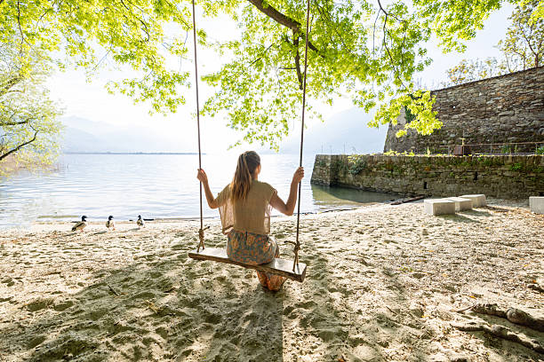 Young woman on wooden swing enjoying tranquil lake view. Peaceful scene in nature with gentle waves. Outdoor relaxation under lush green trees.