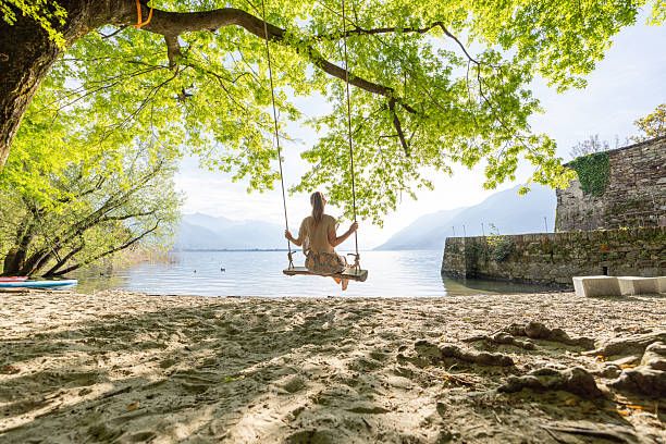 Young woman on wooden swing enjoying tranquil lake view. Peaceful scene in nature with gentle waves. Outdoor relaxation under lush green trees.