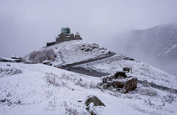 A serene image captures a stone church with a distinctive dome, lightly dusted with snow, nestled on a hillside near Stepantsminda, Georgia. Frost-covered trees stand nearby, adding to the wintry atmosphere. The overcast sky casts a soft light over the quiet, somewhat isolated scene, hinting at the tranquil beauty of the Caucasus region in early winter.