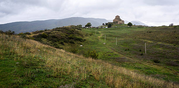 A picturesque view of the historic Jvari Monastery perched atop a hill overlooking the rolling green landscape near Mtskheta, Georgia. The ancient stone structure stands prominently against a backdrop of distant hills and a somewhat overcast sky. A few grazing animals dot the fields below, adding a touch of rural tranquility to the scene.