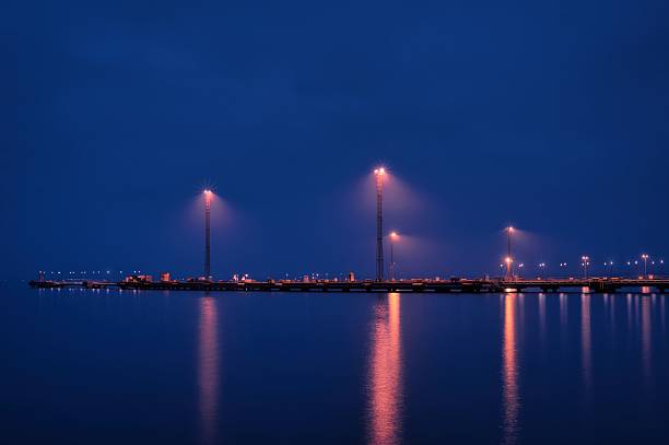 A serene nighttime scene of a harbor illuminated by tall orange streetlights reflecting on the calm surface of the water. The image captures the tranquility of the blue hour with vibrant light reflections and minimalistic composition, making it ideal for themes related to industry, transportation, maritime, and peaceful urban landscapes.