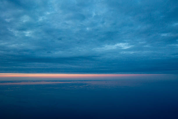 Aerial view above the clouds during sunset. The sky displays a gradient from deep blue at the top to soft pink and orange on the horizon while flying over the Atlantic Ocean and east coast of the USA