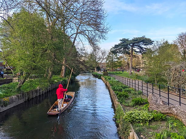 Canterbury, UK - April 19 2025: A traditional punting boat glides along the River Great Stour as people stroll and relax on the tranquil Westgate Gardens banks in this historic English city