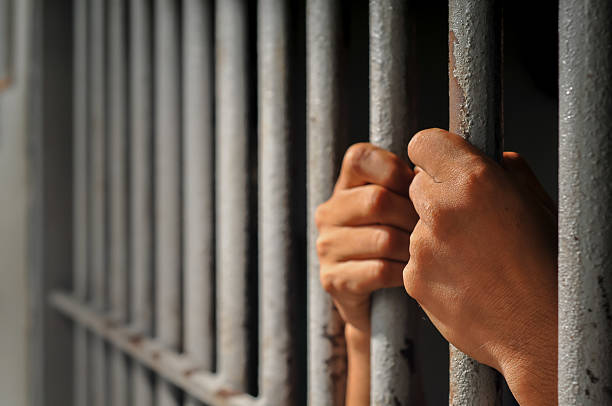istock prisoner's hands behind bars with black background 2213612764