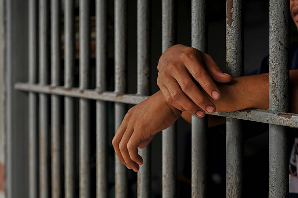 istock prisoner's hands behind bars with black background 2213612765