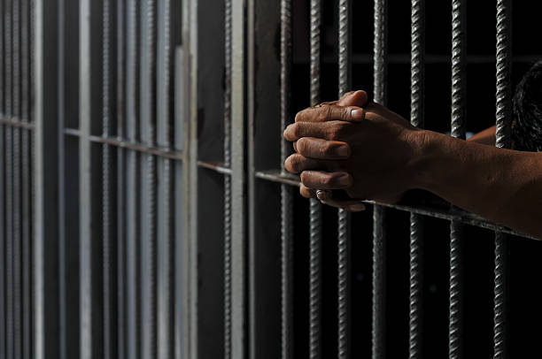 istock prisoner's hands behind bars with black background 2213612837