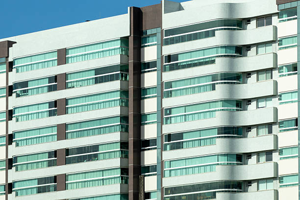 The image shows the facade of a modern building, characterized by striking horizontal lines and large windows with green glass. The architecture combines straight elements with some slightly curved volumes, creating a contemporary look. The predominant colors are white, the brown of the vertical columns and the green of the glass, which together give the building a sophisticated and elegant appearance.