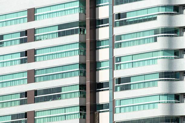 The image shows the facade of a modern building, characterized by striking horizontal lines and large windows with green glass. The architecture combines straight elements with some slightly curved volumes, creating a contemporary look. The predominant colors are white, the brown of the vertical columns and the green of the glass, which together give the building a sophisticated and elegant appearance.