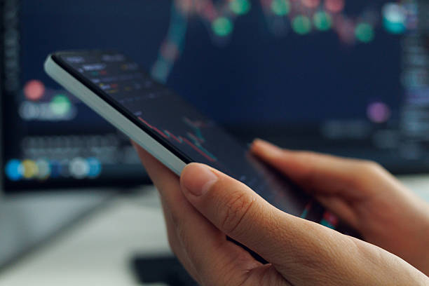 istock A person's hands cradle a smartphone displaying a stock market analysis chart with red and green indicators, set against a softly blurred backdrop of a larger financial data screen, illustrating mobile stock trading or investment monitoring. 2213627043