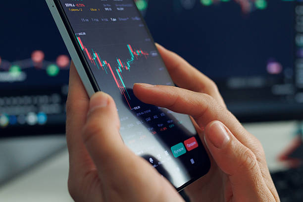 istock A person's hands operate a smartphone displaying a dynamic stock market chart with fluctuating red and green bars, against a blurred background of a larger monitor showing similar financial data, representing mobile trading or investment management. 2213627054