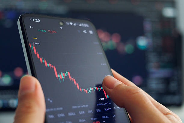 istock A person's hands hold a smartphone displaying a declining stock market chart with mostly red candlesticks, indicating a negative trend, against a blurred backdrop of a larger financial data screen, illustrating mobile trading during a market downturn. 2213627057