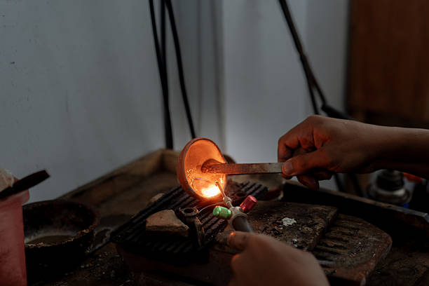 An artisan uses a handheld torch to melt metal in a small clay crucible during a traditional casting process. The focused flame and rustic workspace highlight the skilled, hands-on nature of metal craftsmanship.