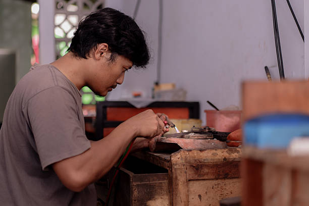 A jewelry artisan carefully soldering silver with a torch, intense heat glowing as flame meets metal. Beside the flame, a silver ring sits on a firebrick, awaiting its next transformation.