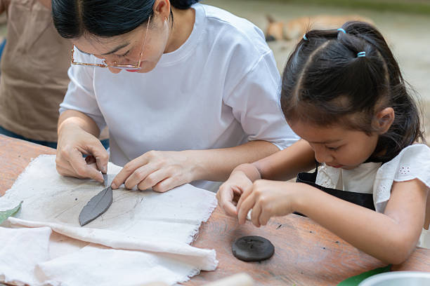 A mother and daughter work together with clay on a table, possibly crafting or sculpting.