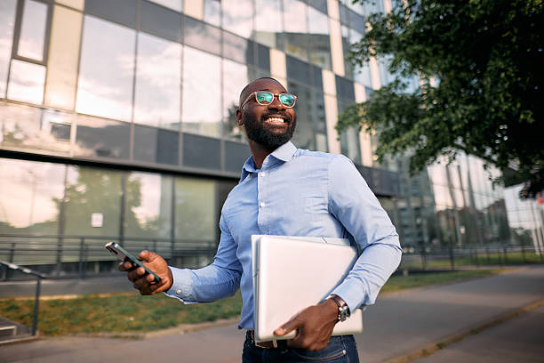 istock Smiling businessman walking outside office using smartphone and holding laptop 2213659335