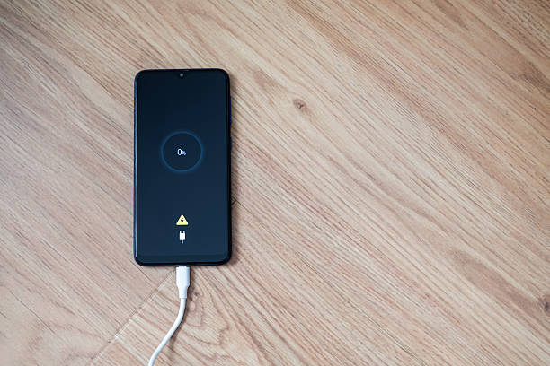 istock White charging cable plugged into smartphone displaying 0% battery alongside a low battery warning symbol, resting on a wooden table with a clean, minimal surface 2213664293
