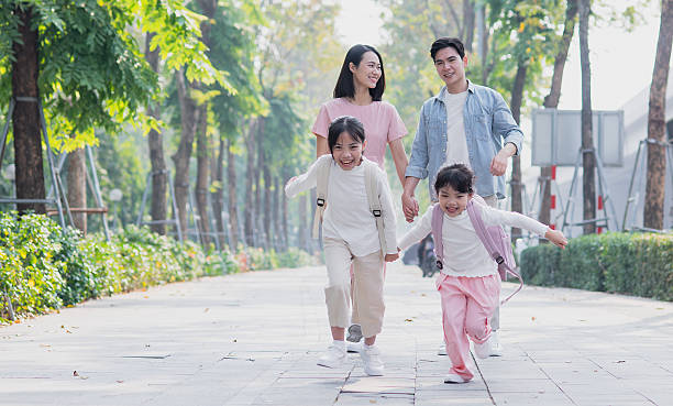 A family walks hand in hand on a sunny day, with two joyful children running ahead. Trees line the pathway, creating a warm and inviting atmosphere.