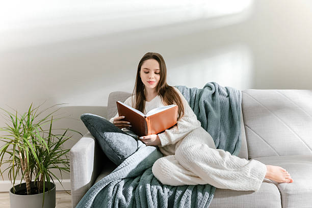 istock Young woman in soft loungewear reading a book while relaxing on a light sofa in natural light. 2213673414