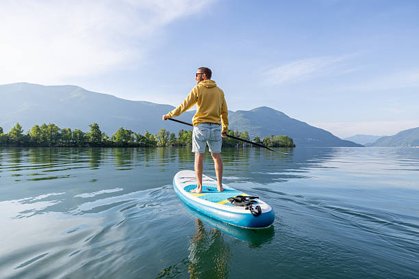 Man in yellow hoodie enjoys paddleboarding on a calm lake. Scenic mountain backdrop enhances the peaceful outdoor adventure. Ideal for leisure, travel, and nature themes.